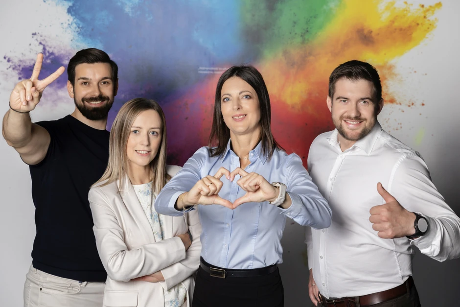 Cztery uśmiechnięte osoby pozują na tle kolorowej ściany, tworząc pozytywne gesty dłoni.