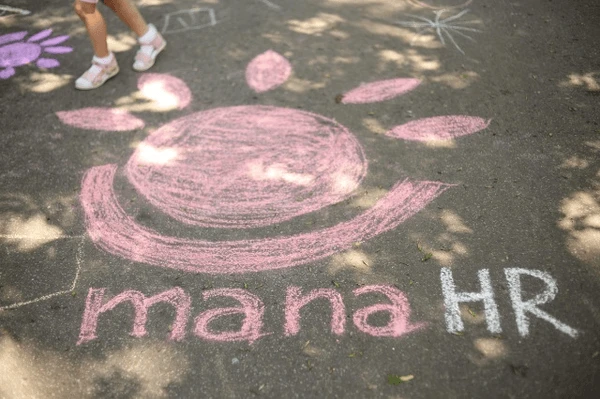 Radosny różowy rysunek słońca i napisu mana HR namalowany kredą na asfalcie w cieniu drzew