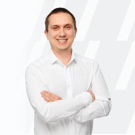 Mężczyzna w białej koszuli z założonymi rękami uśmiecha się na jasnym tle z abstrakcyjnym wzorem