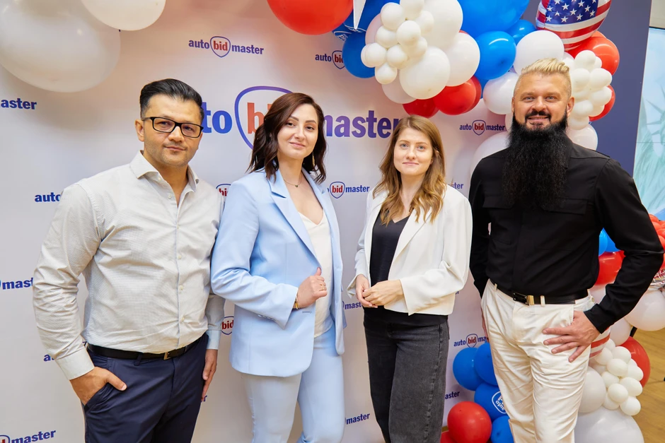 Zespół pracowników firmy AutoBidMaster pozujący na tle firmowego baneru i kolorowych balonów