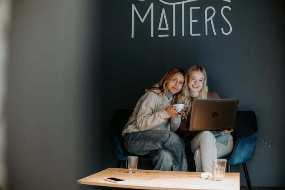 Dwie kobiety siedzą razem na fotelach, pracując na laptopie i pijąc kawę w przytulnym biurze