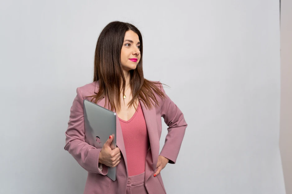 Młoda kobieta w różowym garniturze trzyma laptop i patrzy w bok na jasnym tle