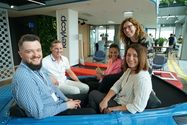 Pięć uśmiechniętych osób siedzi w niebieskim samochodzie w nowoczesnym biurze firmy Keyloop