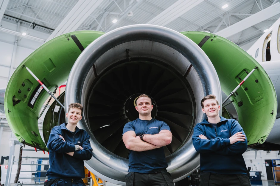 Trzech młodych mechaników stoi z założonymi rękami przed silnikiem odrzutowego samolotu w hangarze lotniczym