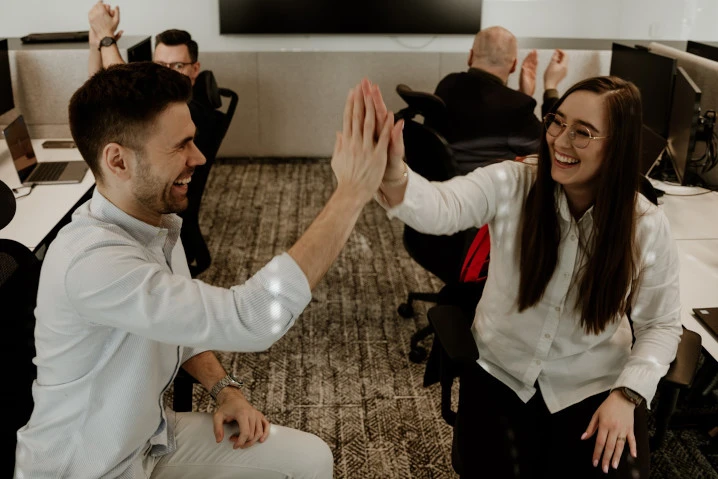 Dwóch uśmiechniętych pracowników firmy daje sobie piątkę w nowoczesnym biurze przy stanowiskach pracy