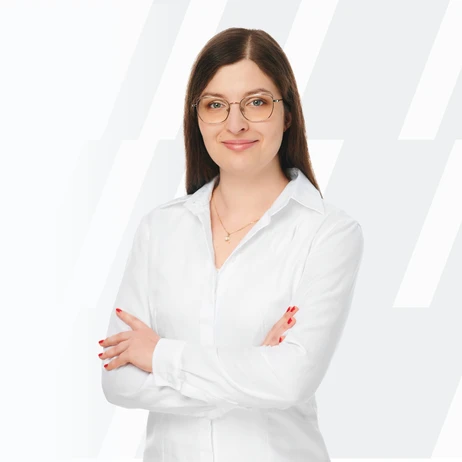 Młoda kobieta z okularami w białej koszuli stoi z założonymi rękami na jasnym tle