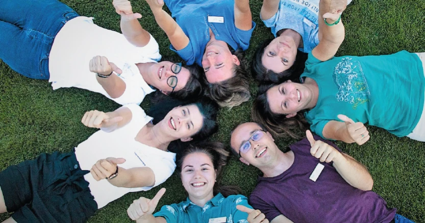 Zespół siedmiu osób leżących na trawie w kółku, uśmiechnięci z kciukami uniesionymi do góry