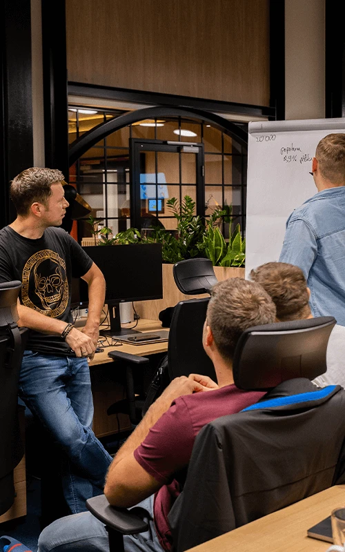 Zespół pracowników omawia projekt przy tablicy w nowoczesnym biurze z roślinami i komputerami