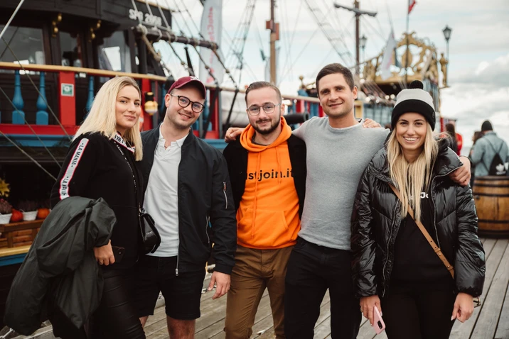 Grupa młodych ludzi pozująca razem na drewnianym nabrzeżu z kolorowym statkiem w tle