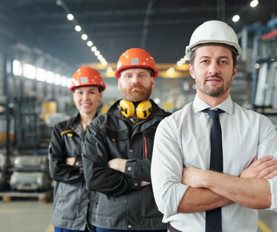 Zespół pracowników w kaskach ochronnych w hali produkcyjnej stojący z założonymi rękami