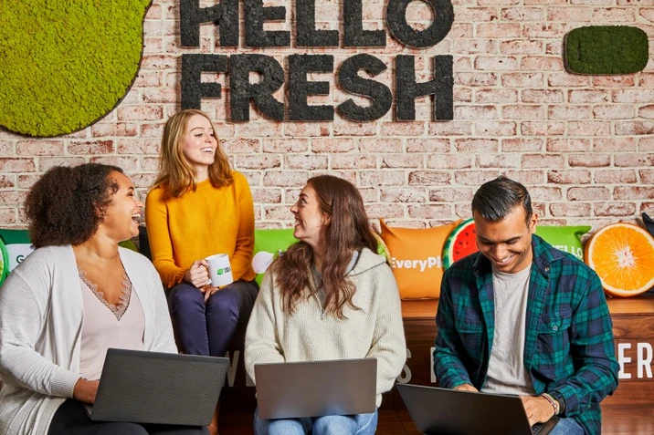 Cztery osoby w biurze Hello Fresh pracują i rozmawiają w przyjaznej atmosferze przy laptopach