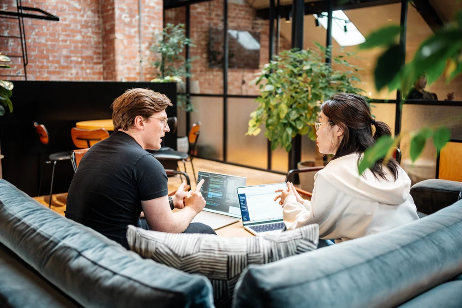 Dwóch pracowników omawia projekt przy laptopach w nowoczesnym biurze z ceglaną ścianą i roślinami