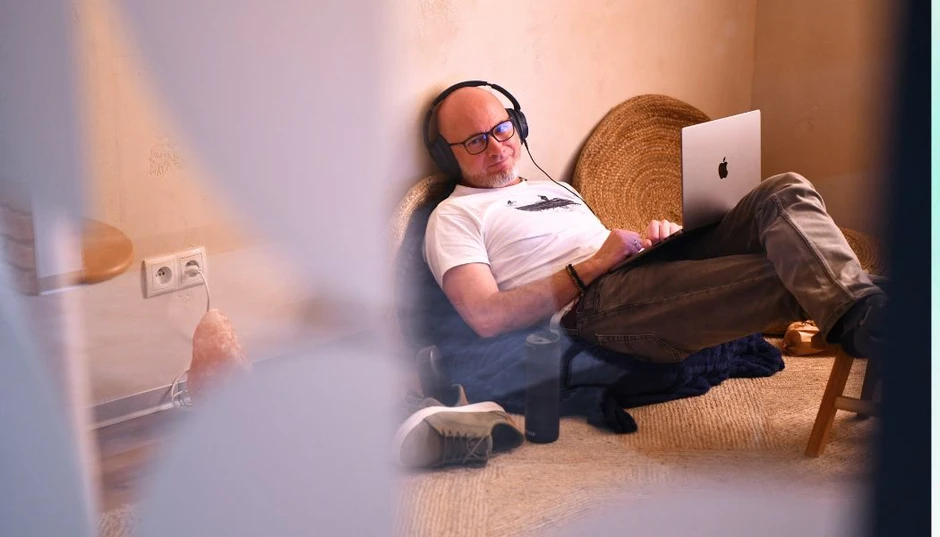 Mężczyzna w okularach i słuchawkach pracuje na laptopie siedząc na podłodze w przytulnym kąciku biurowym