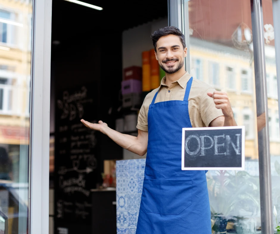 Uśmiechnięty mężczyzna w fartuchu zaprasza do otwartego sklepu lub kawiarni w miejskim otoczeniu