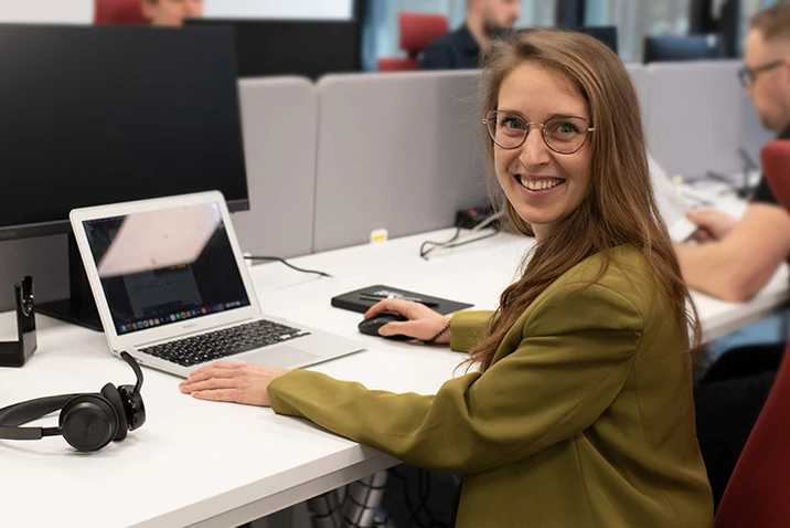 Młoda kobieta uśmiechająca się przy laptopie w nowoczesnym biurze open space podczas pracy zespołowej