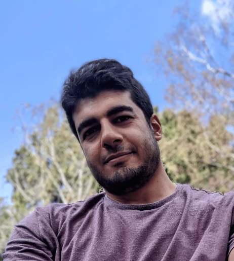 Zdjęcie przedstawia mężczyznę w jasnofioletowej koszulce na tle nieba i zielonych drzew podczas słonecznego dnia