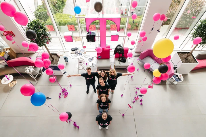 Grupa pracowników w czarnych koszulkach w biurze z różowymi i kolorowymi balonami oraz dużym różowym logo