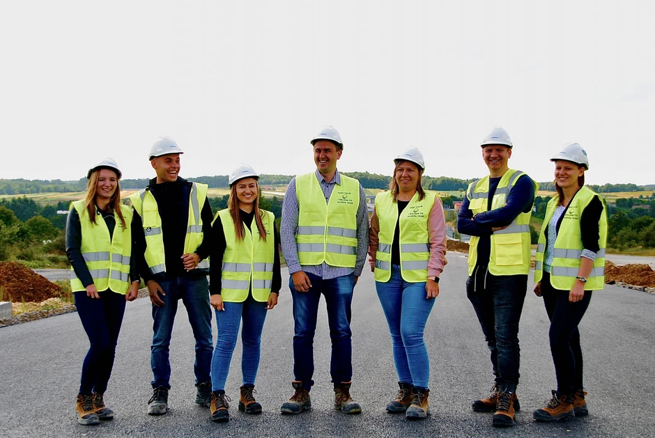 Zespół pracowników budowlanych w kaskach i kamizelkach odblaskowych stoi razem na placu budowy na zewnątrz