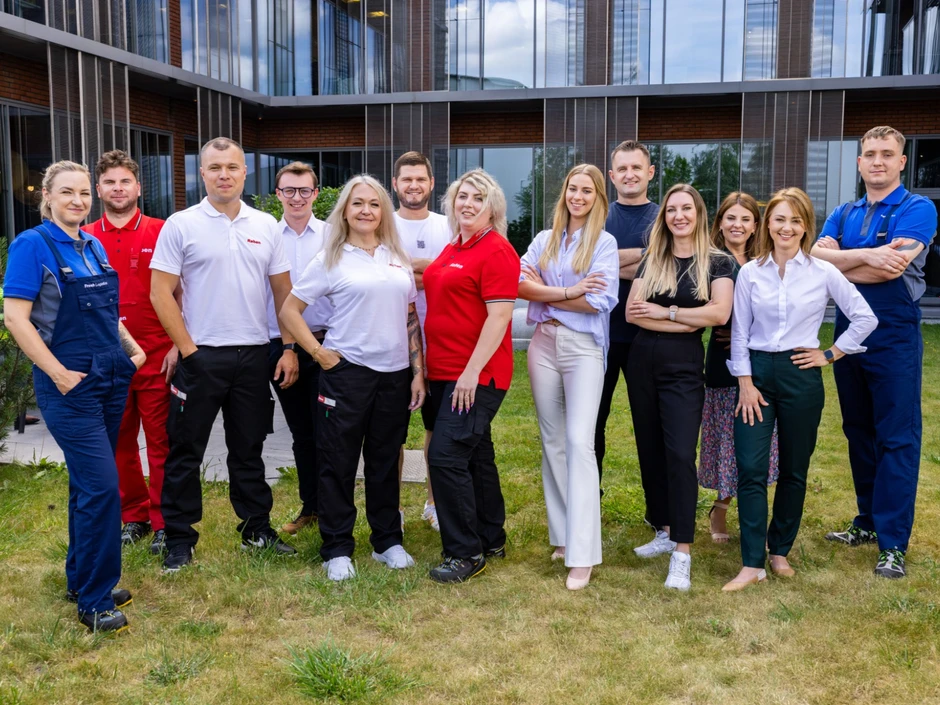 Zespół pracowników w różnorodnych strojach firmowych stoi razem na trawie przed nowoczesnym budynkiem biurowym