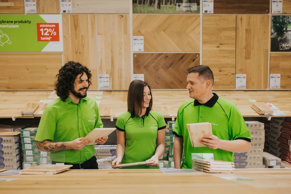Trójka pracowników w zielonych koszulkach omawia i prezentuje drewniane panele podłogowe w sklepie