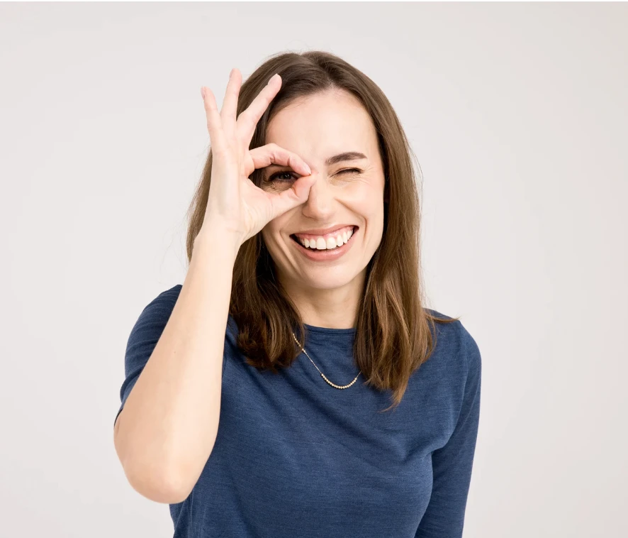 Uśmiechnięta kobieta pokazuje gest OK jednym okiem patrząc przez utworzony palcami okrąg