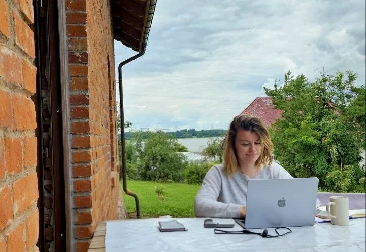 Kobieta pracująca na laptopie na zewnątrz przy stole z widokiem na zieleń i wodę