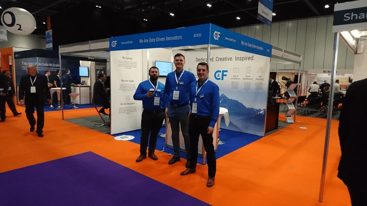 Trzej mężczyźni w niebieskich koszulach pozują na stoisku firmy na targach branżowych z motywem danych