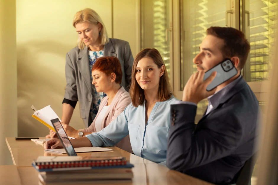 Cztery osoby pracujące w biurze, jedna kobieta uśmiechnięta przy laptopie, mężczyzna rozmawia przez telefon