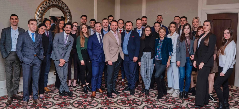 Grupa 28 osób w różnym wieku pozuje na tle jasnej ściany i ozdobnego dywanu w sali konferencyjnej