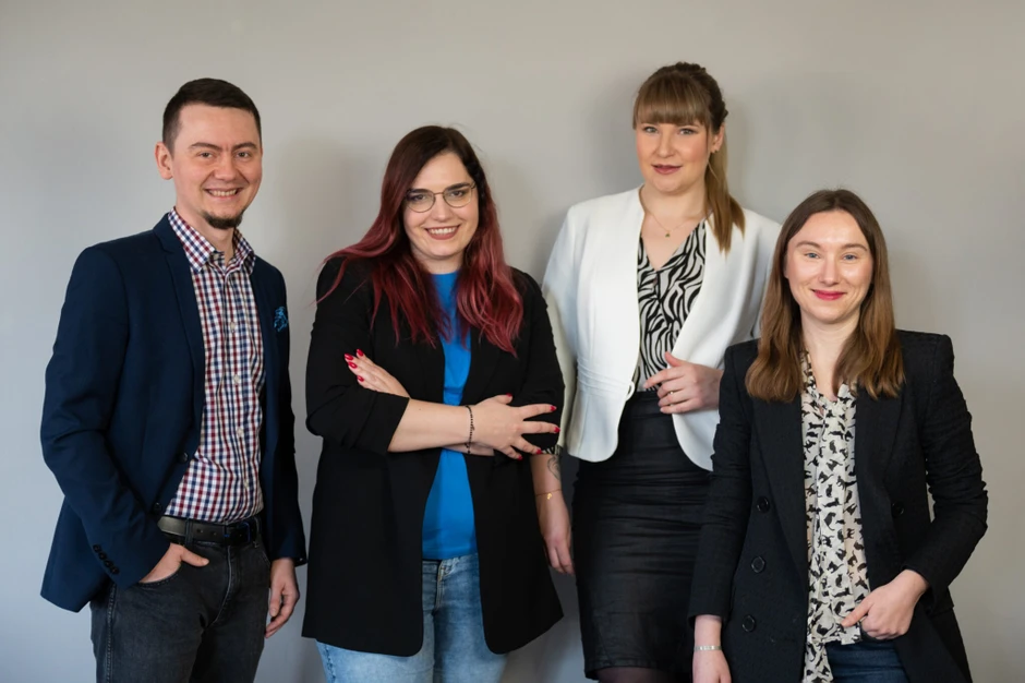 Cztery osoby w biznesowym stroju pozują uśmiechnięte na tle szarej ściany w nowoczesnym biurze