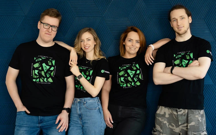 Cztery osoby w czarnych koszulkach z grafiką stoją na tle niebieskiej ściany, uśmiechnięte i zrelaksowane