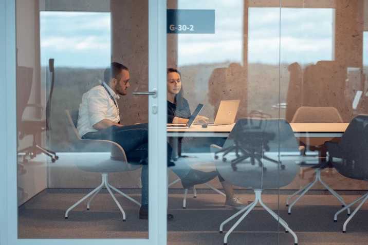 Dwóch pracowników omawia projekt przy laptopach w nowoczesnej sali konferencyjnej z przeszklonymi ścianami