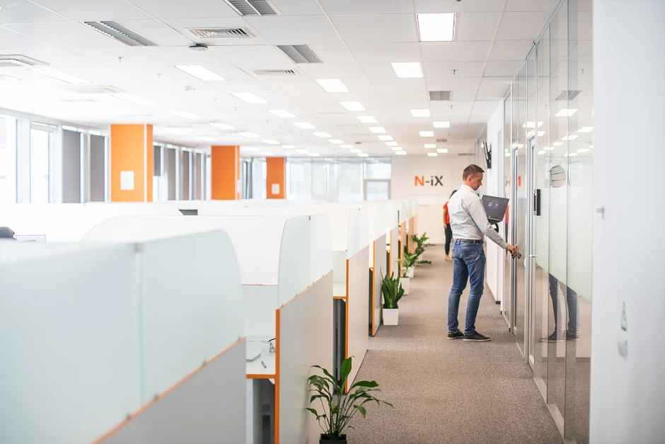 Nowoczesne biuro z białymi ściankami działowymi, roślinami i pracownikiem otwierającym szklone drzwi