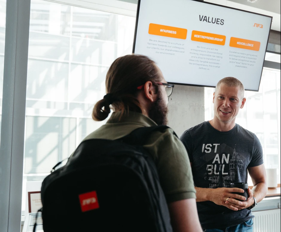 Dwóch mężczyzn rozmawia w nowoczesnym biurze z ekranem prezentującym wartości firmy w tle