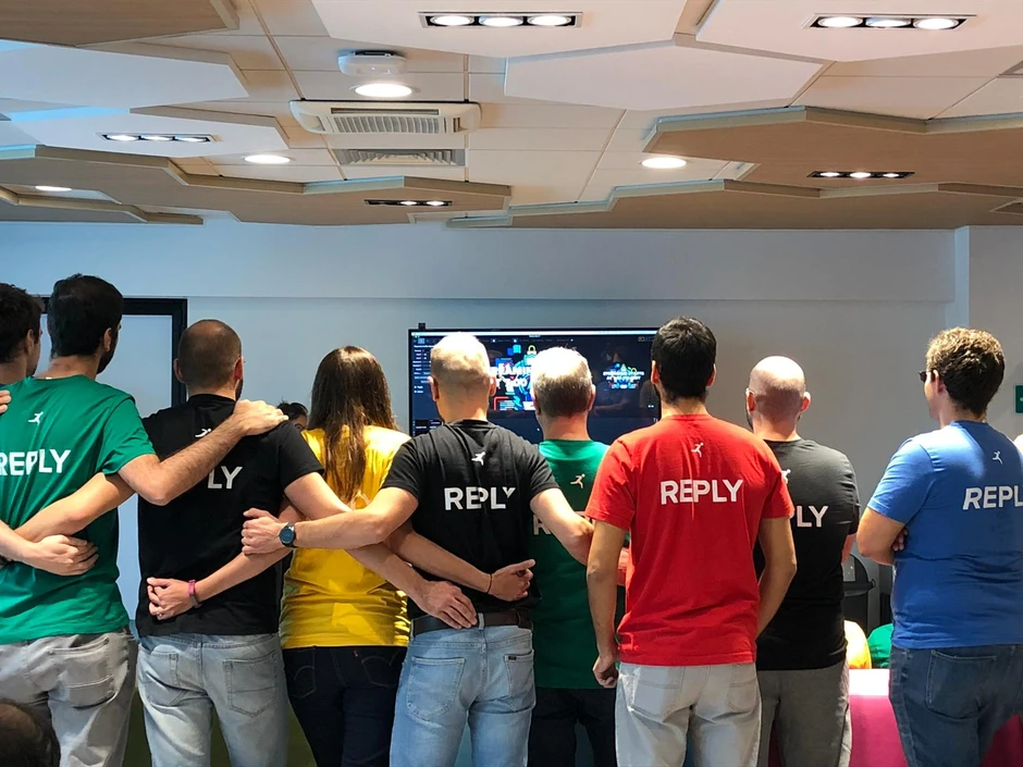 Grupa pracowników w różnokolorowych koszulkach Reply stoi razem w biurze z połączonymi ramionami