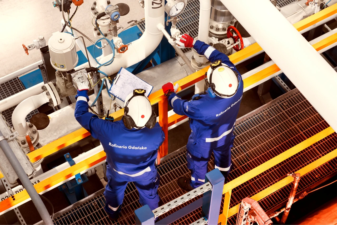 Dwóch pracowników Rafinerii Gdańskiej w odzieży ochronnej kontroluje urządzenia przemysłowe i analizuje dokumentację