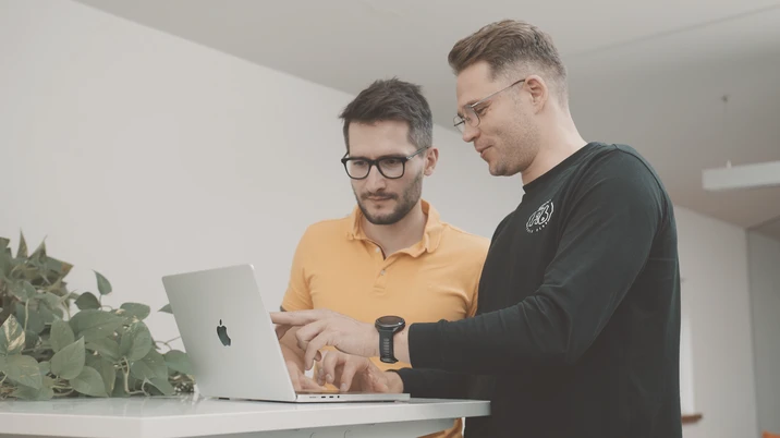 Dwóch mężczyzn pracuje wspólnie przy laptopie Apple w nowoczesnym biurze z roślinami w tle