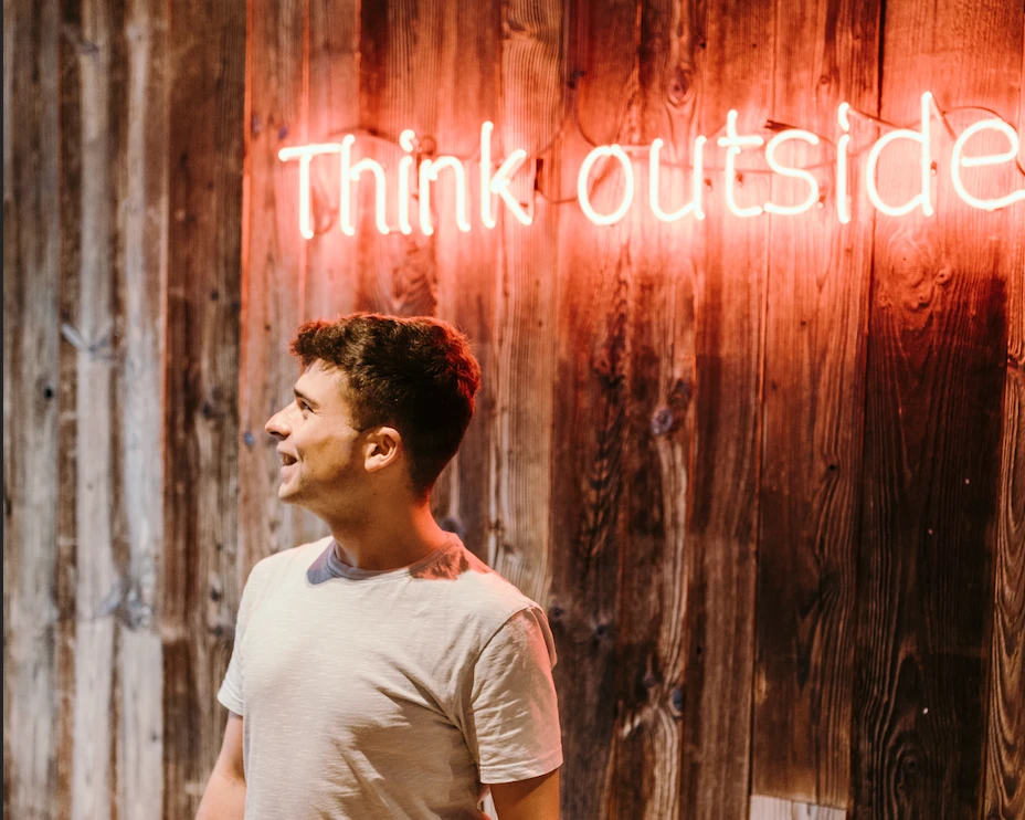 Młody mężczyzna stoi z uśmiechem przed drewnianą ścianą z napisem neonowym Think outside