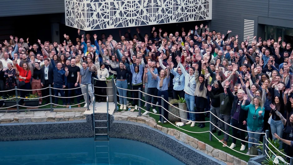 Duża grupa pracowników pozująca razem na tle biura obok basenu, wszyscy pozdrawiają kamerę