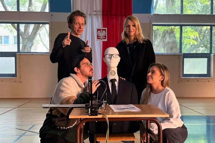 Cztery osoby wokół automatycznego manekina w garniturze siedzącego przy biurku w jasnej hali sportowej