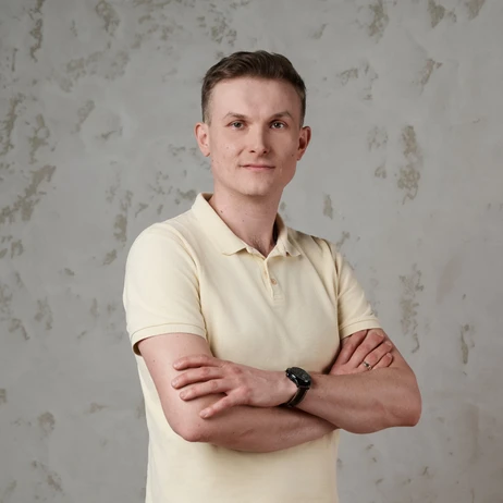 Mężczyzna w jasnej koszulce polo stoi pewnie z założonymi rękami na tle jasnej ściany z delikatnymi wzorami