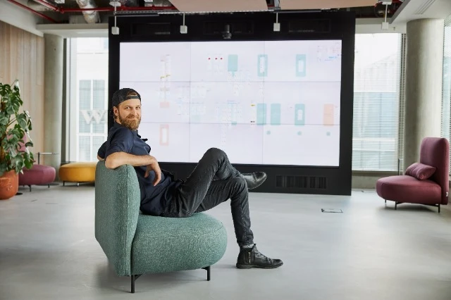 Mężczyzna w czapce siedzi na zielonym fotelu w nowoczesnym biurze z dużym ekranem na ścianie