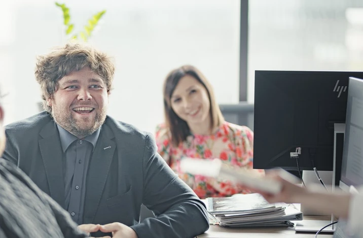 Zespół pracowników w biurze podczas spotkania, na pierwszym planie uśmiechnięty mężczyzna w garniturze