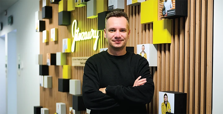 Uśmiechnięty mężczyzna z założonymi rękami stoi na tle nowoczesnej ściany z logo firmy i zdjęciami pracowników