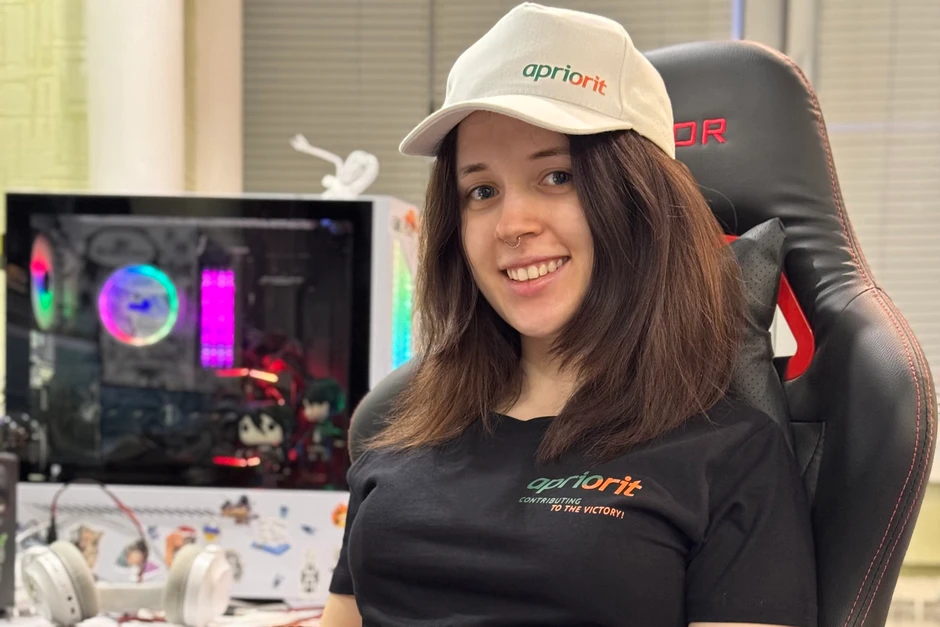 Młoda kobieta w czarnej koszulce i czapce z logo Apriorit siedzi w fotelu gamingowym w biurze
