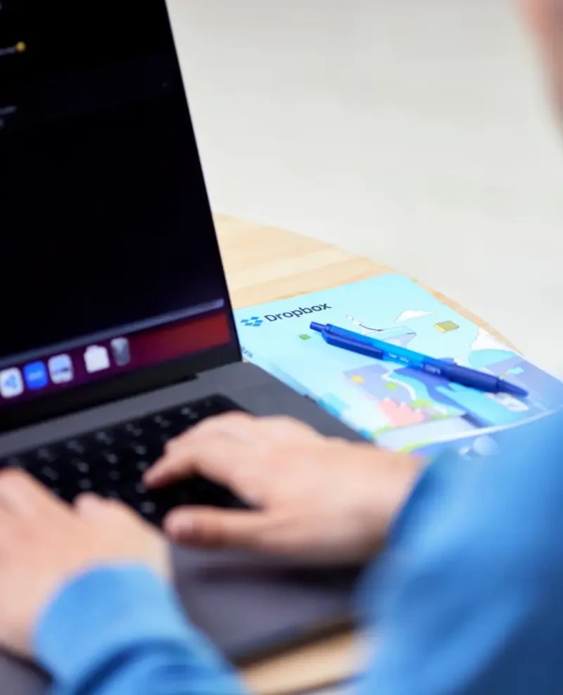 Osoba pracująca na laptopie obok notesu z logo Dropbox i długopisu na drewnianym biurku