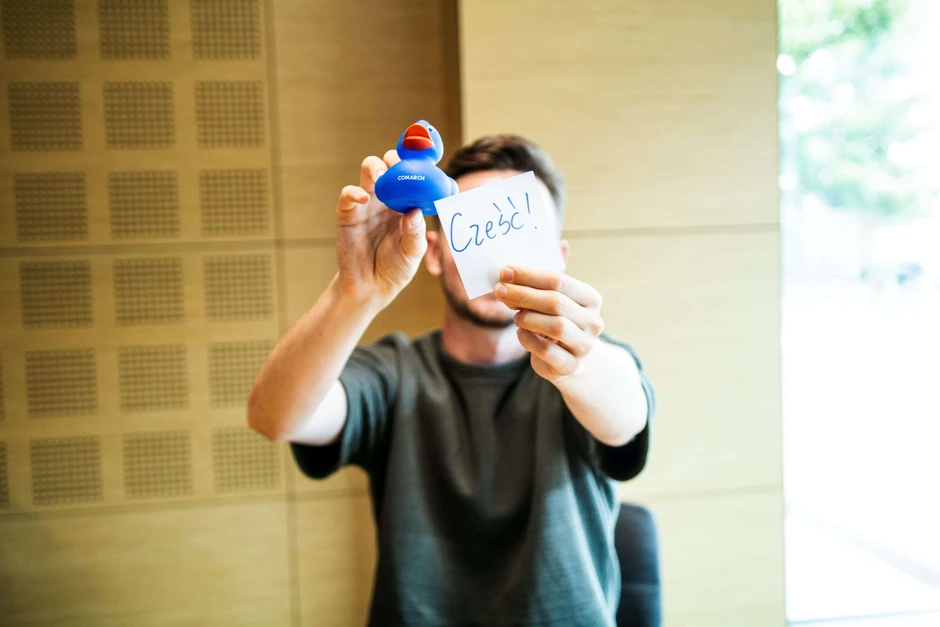 Mężczyzna siedzi w nowoczesnym biurze trzymając niebieską gumową kaczkę z napisem Comarch i kartkę z napisem Cześć