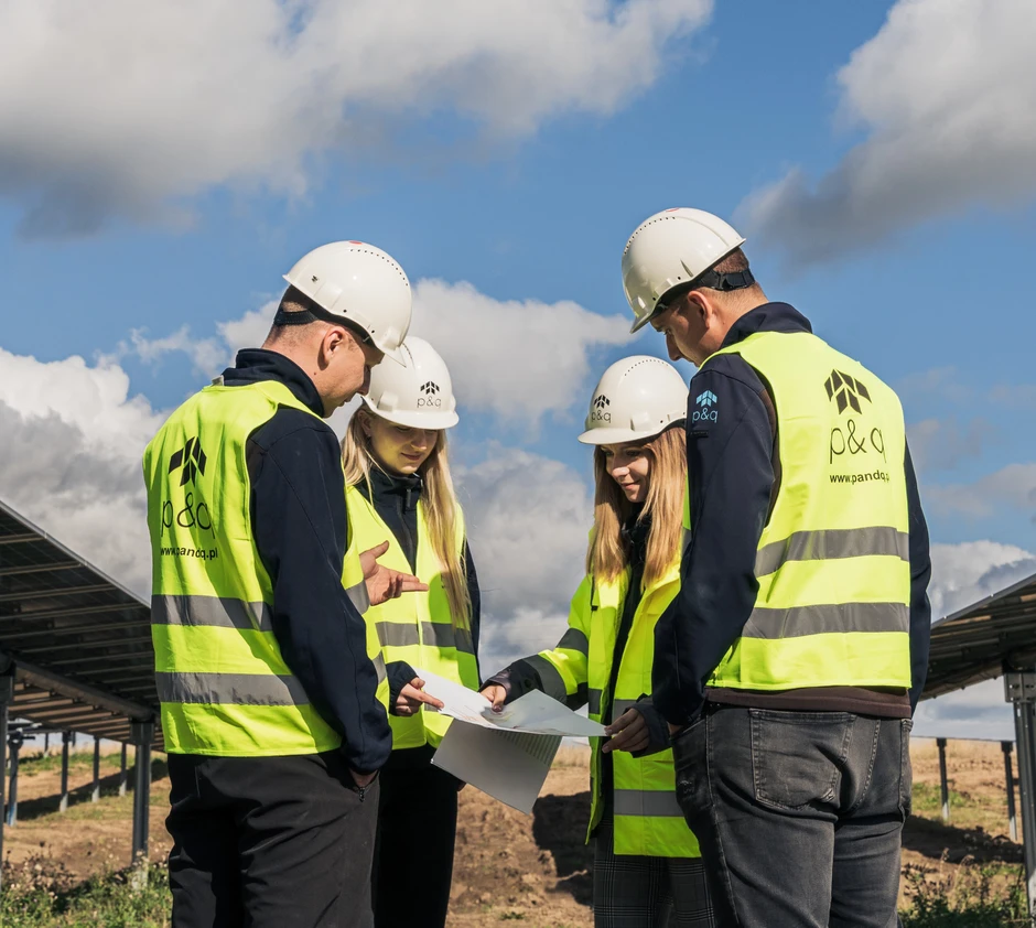 Czterech pracowników w kamizelkach i kaskach omawia projekt przy panelach słonecznych na zewnątrz