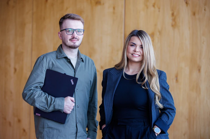 Dwójka młodych pracowników w biurze na tle drewnianej ściany pozują do profesjonalnego zdjęcia firmowego