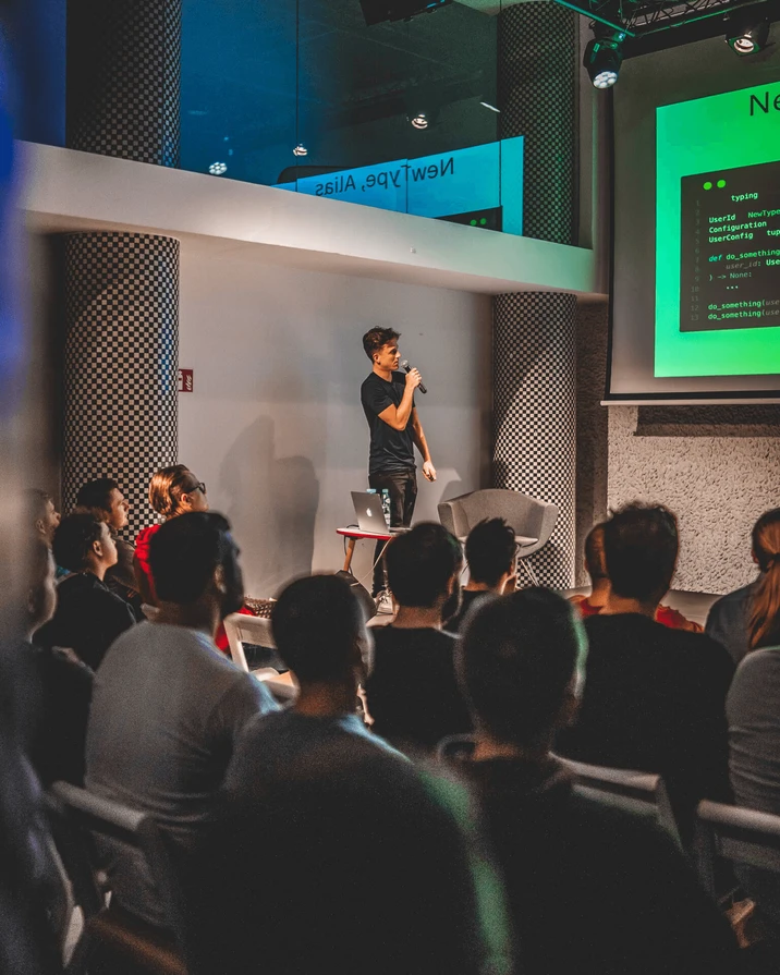 Młody mężczyzna prowadzi prezentację techniczną przed zgromadzonymi słuchaczami w nowoczesnej sali konferencyjnej